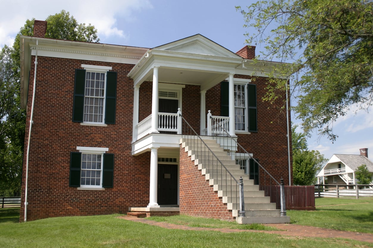 A historic East Coast brick building with white columns, black shutters, and a prominent staircase leading to the second-floor entrance, surrounded by green grass and trees on a sunny day.