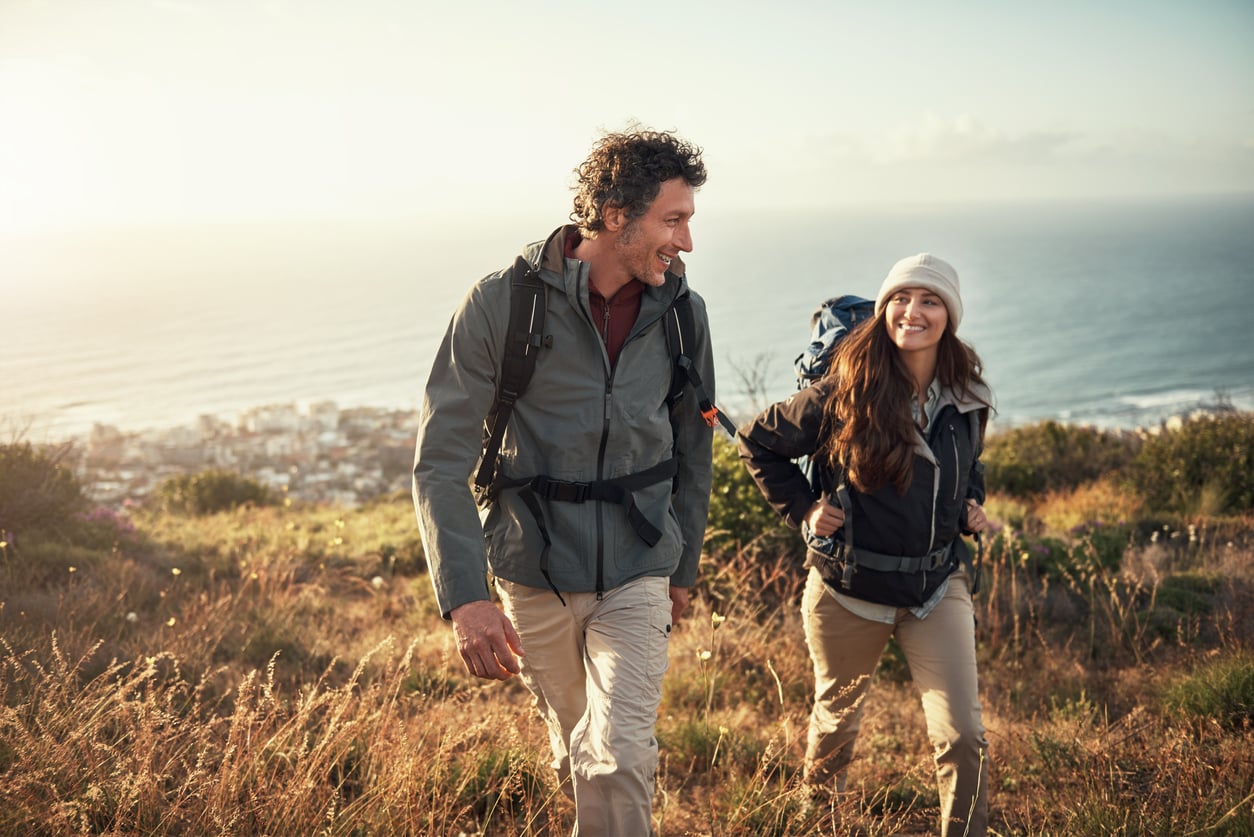 A man and woman with backpacks hike up a grassy hill overlooking the ocean and a coastal town, smiling at each other under a clear sky, enjoying the simple pleasures of adventure fueled by their money saving habits.