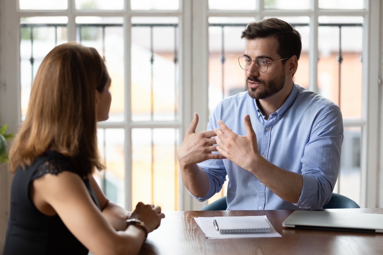 A man with glasses gestures while talking to a woman across a table in a bright office, sharing his Baby Boomer beliefs. The woman listens attentively, and a notebook and laptop are on the table. Large windows are in the background.