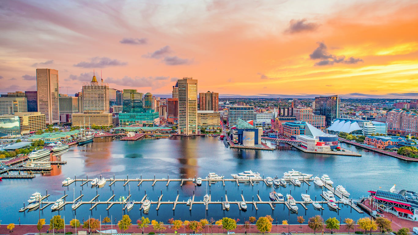 Aerial view of Baltimore’s Inner Harbor at sunset, with boats docked in the marina and tall buildings—hallmarks of one of the big cities in America—reflecting colorful sky clouds on the water.
