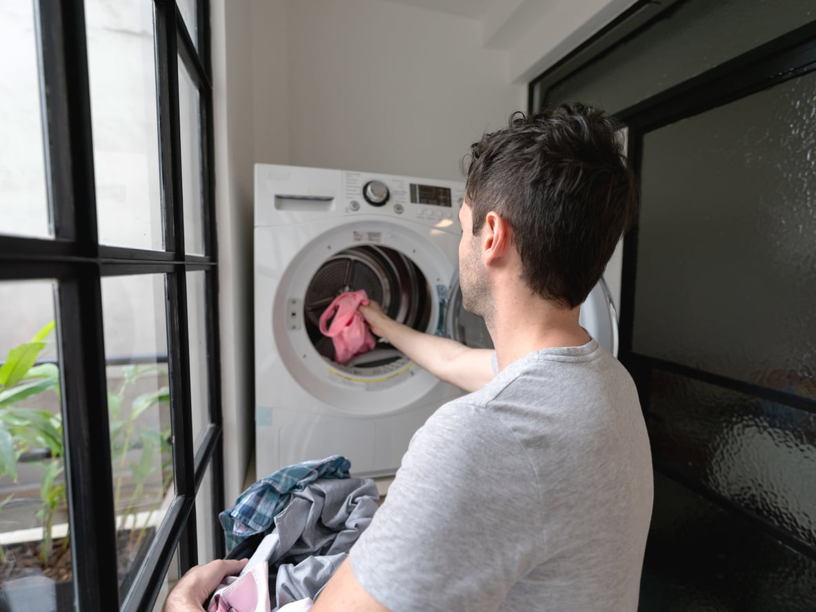 A man in a gray t-shirt loads colorful laundry into a front-loading washing machine in a bright laundry room with large windows.