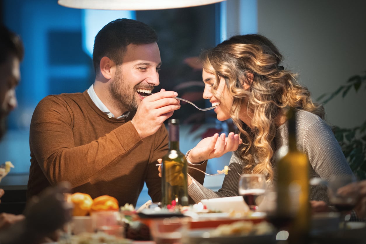 A man smiles as he feeds a woman with a fork at a cozy dinner table set with wine, food, and candles. Both look happy and are enjoying their meal together.