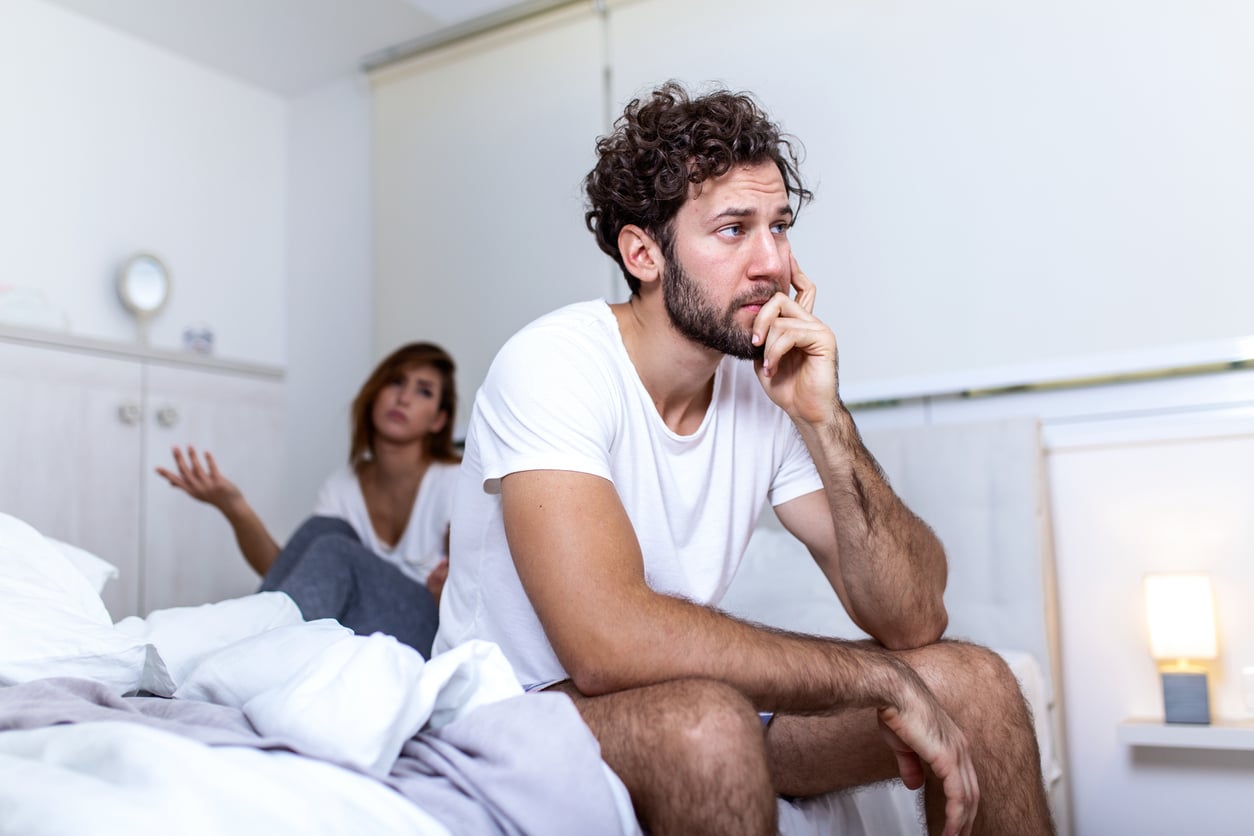A man sits on a bed looking worried while a woman sits behind him gesturing with her hands, suggesting a disagreement or emotional conversation in a bright bedroom.