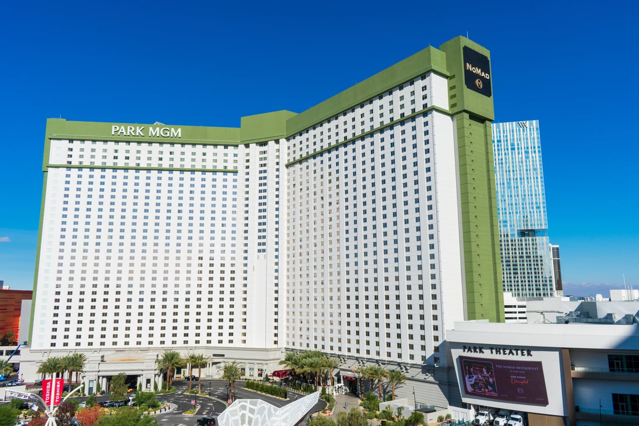 Park MGM hotel in Las Vegas, a tall white building with green accents, stands against a clear blue sky. The Park Theater entrance and palm trees are visible in front.