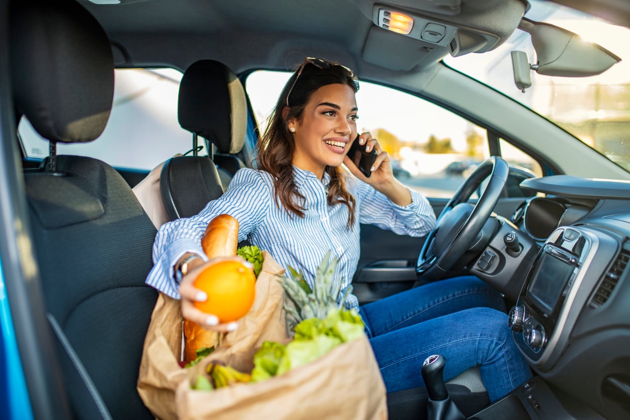 A smiling woman sits in a car, talking on her phone while holding a paper bag full of groceries, including a baguette and an orange, on her lap—perhaps swapping car maintenance hacks with a friend.