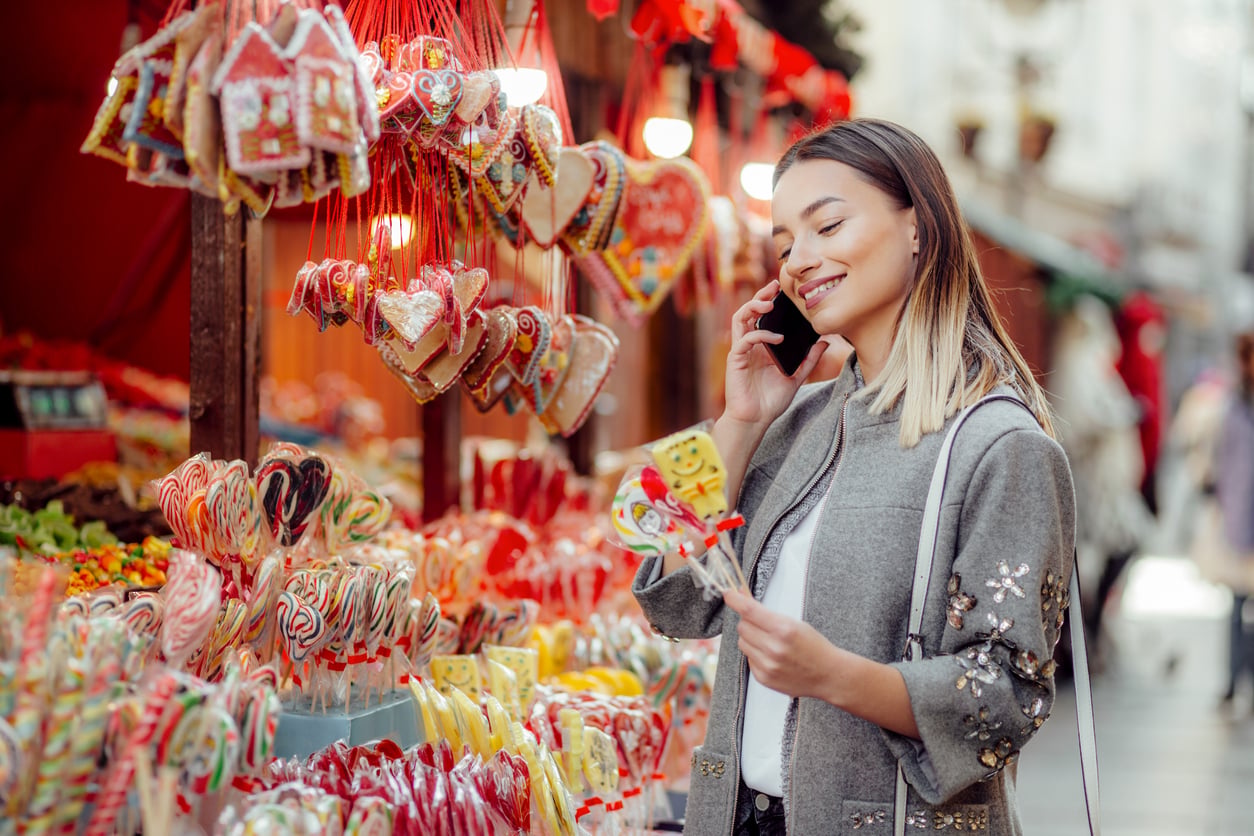 A woman smiles while talking on the phone, standing at a colorful outdoor market stall filled with various candies and heart-shaped sweets.