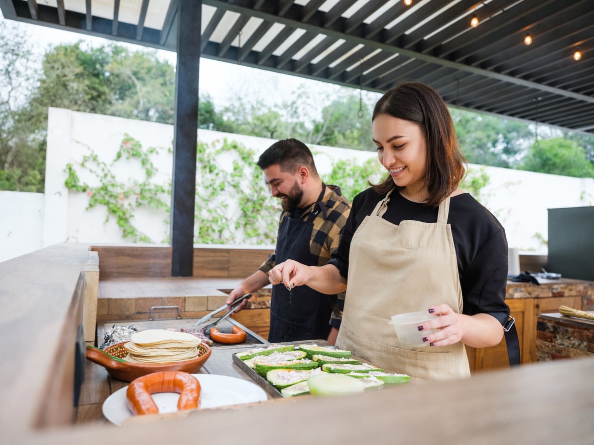 Two people are cooking outdoors under a pergola, enjoying the comfort of quality outdoor home products. The woman in front is spreading something on sliced vegetables, while the man in the background grills sausages. Both are wearing aprons and smiling.