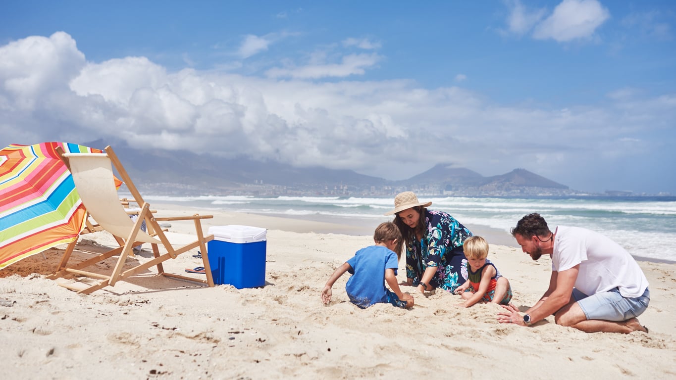 A family of four, with two children and two adults, enjoys quality time playing in the sand on a sunny beach. Their beach vacation savings made this special day possible, as they relax by a colorful umbrella, beach chair, and blue cooler beneath the mountains.