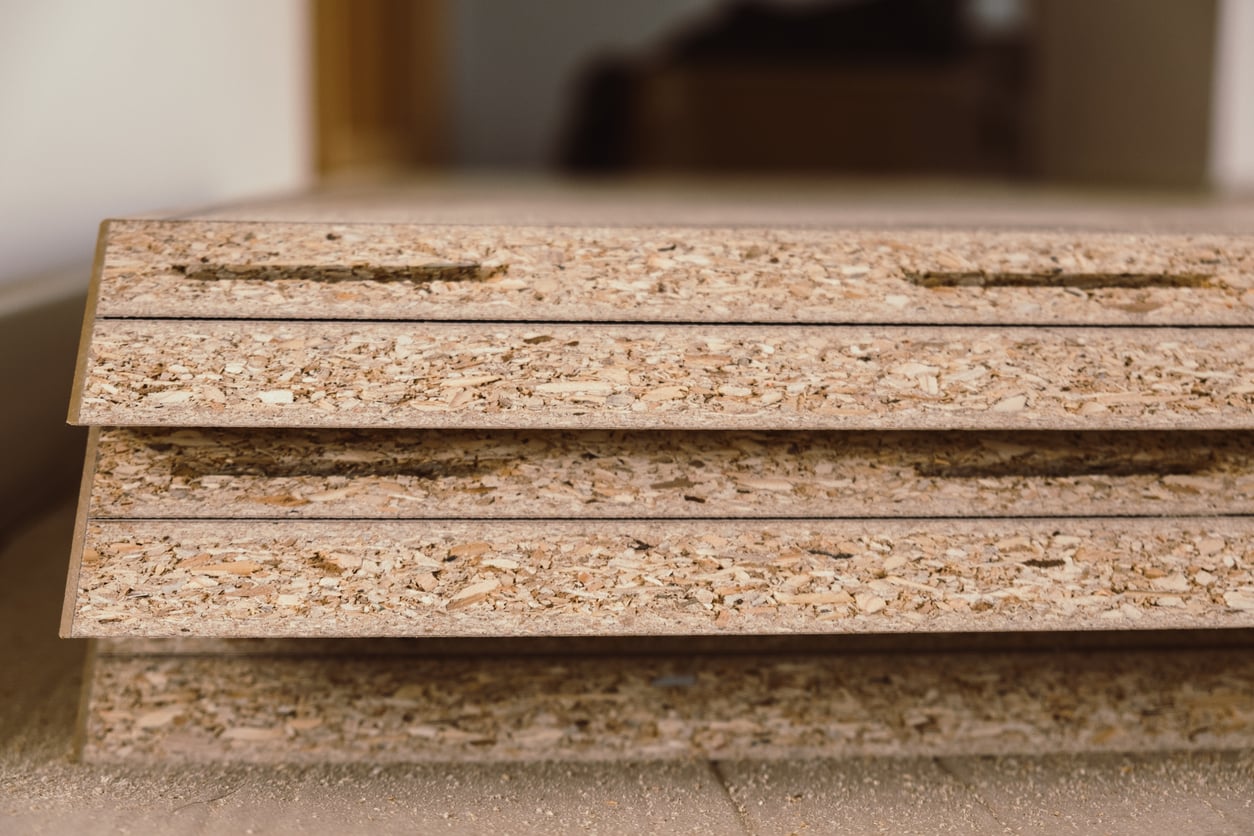 Close-up of stacked particle board sheets with grooves, resting on a floor indoors. The texture and compressed wood chips of each sheet are clearly visible, illustrating why some consider them to be waste of money products due to their lower durability compared to solid wood.