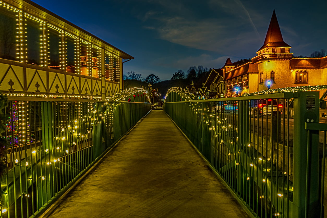 A pedestrian bridge decorated with string lights leads to buildings adorned with festive lights at night, creating a warm, inviting holiday atmosphere reminiscent of the best Christmas towns in America.