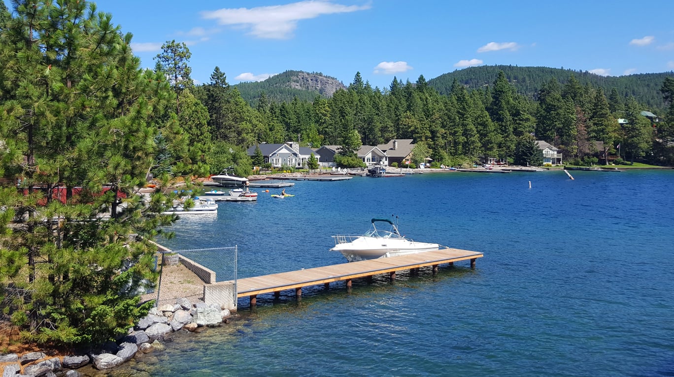 A small white boat docked on a wooden pier extends into a blue lake, surrounded by green trees, lakeside houses, and forested hills under a clear, sunny sky.