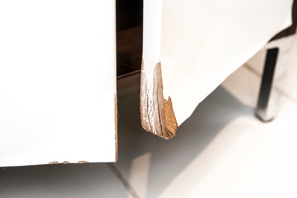 Close-up of a white cabinet door with a large, chipped and peeling bottom corner, exposing the brown wood underneath—a clear example of products that don't last. The cabinet stands on a tiled floor.