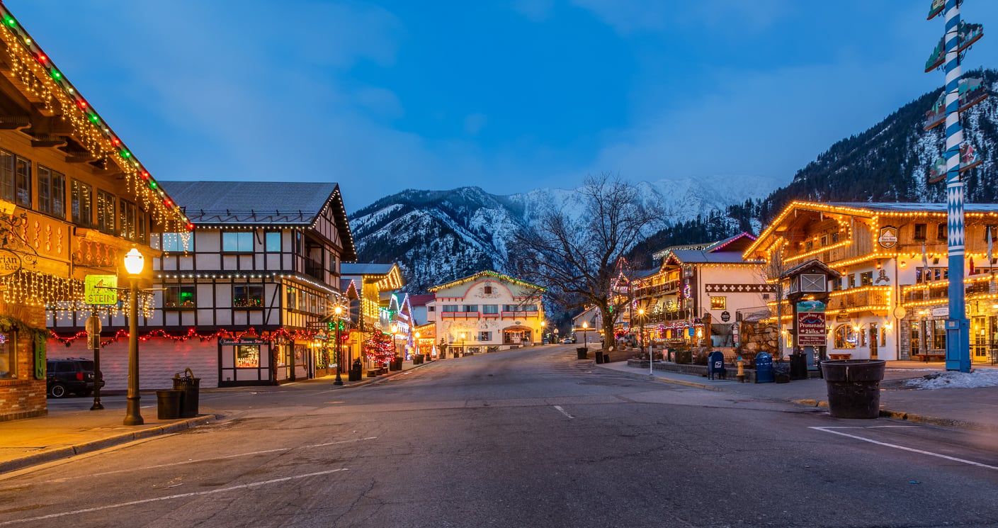 A picturesque street lined with Bavarian-style buildings adorned with colorful holiday lights, set against snow-capped mountains at dusk, creates a festive, winter atmosphere reminiscent of the best Christmas towns in America.