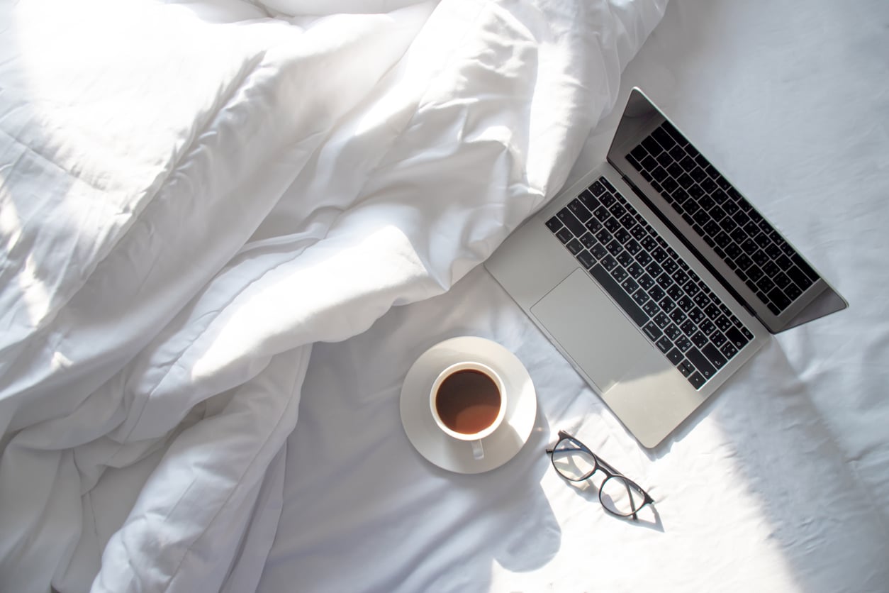 A cup of coffee, a laptop, and a pair of glasses rest on a white bed with rumpled sheets, bathed in soft natural light.