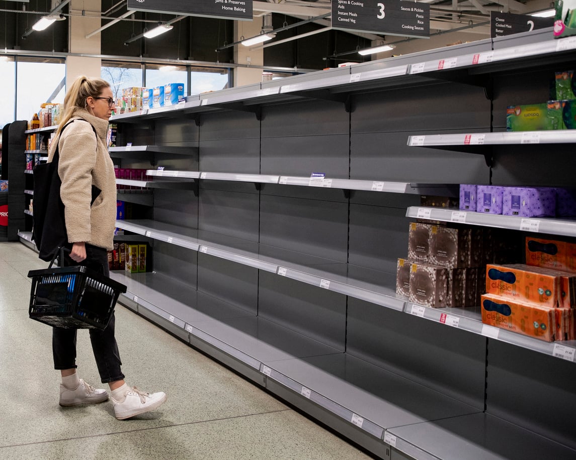 A woman with a shopping basket stands in front of nearly empty supermarket shelves, looking at the limited selection of items left.