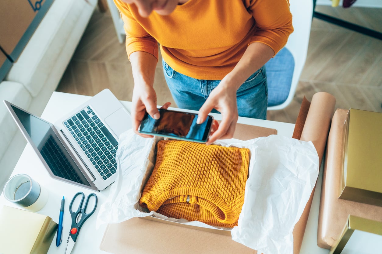 A person in a yellow sweater takes a photo of a folded yellow knit sweater in a shipping box, with a laptop, scissors, tape, pen, and wrapped packages on the nearby white desk—a scene that captures how women are changing their spending behaviors in today’s evolving marketplace.