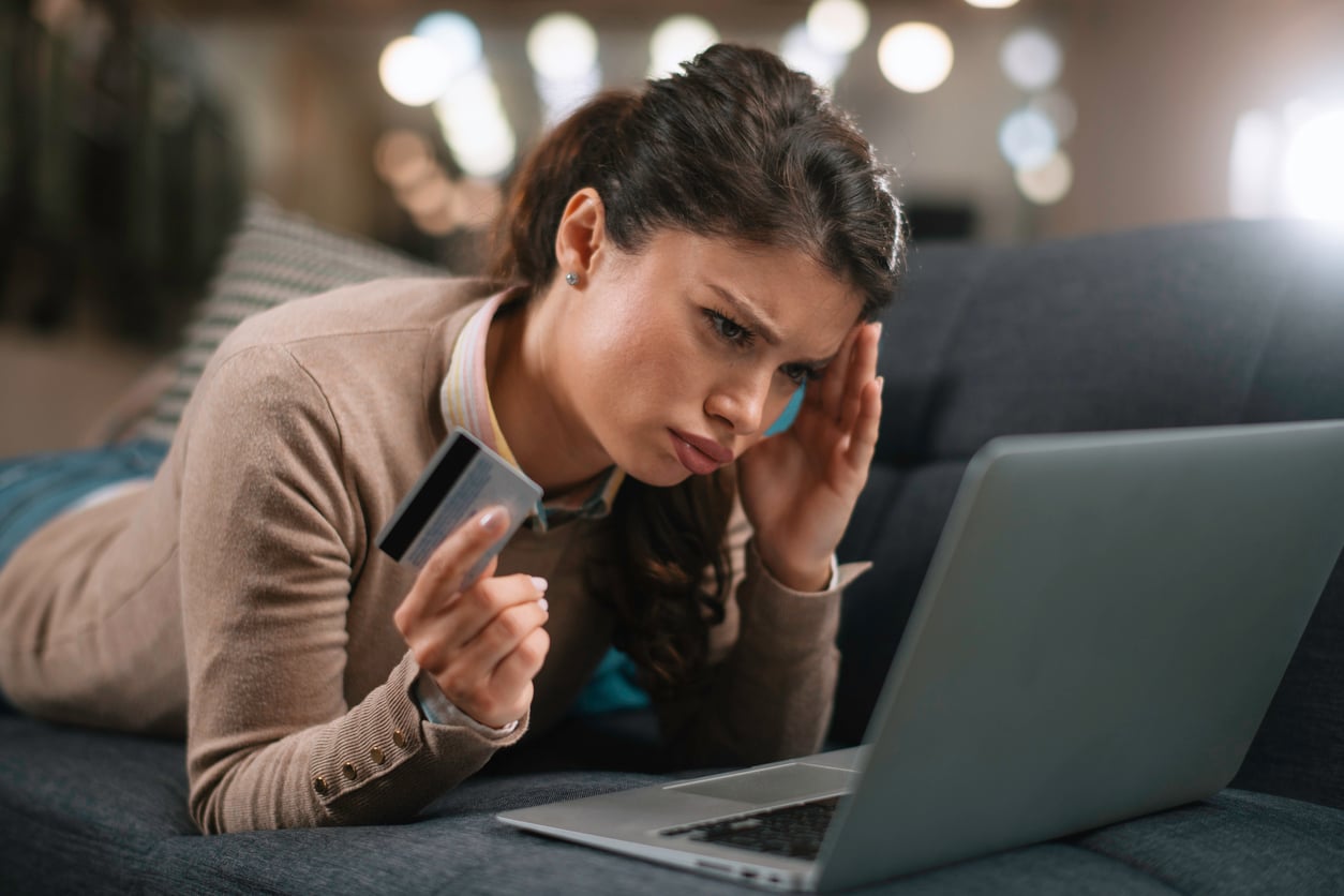 A woman lies on a couch holding a credit card and looking worriedly at a laptop screen, reflecting how women are changing their spending behaviors amid uncertainty with online shopping or banking.