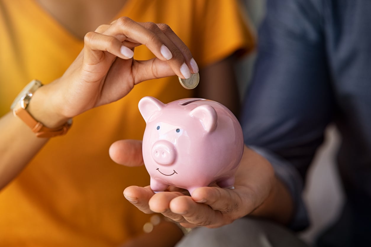 A person drops a coin into a pink piggy bank held by another person. Both are wearing casual clothes, and only their hands and part of their torsos are visible. The scene suggests saving money or financial planning.