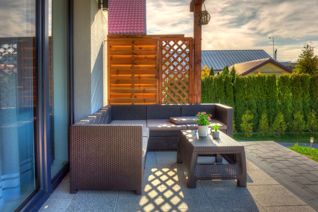 Modern patio with wicker furniture, grey cushions, a coffee table with potted plants, and a wooden privacy screen. Sunlight casts shadows on the tiled floor, and a green hedge lines the background.