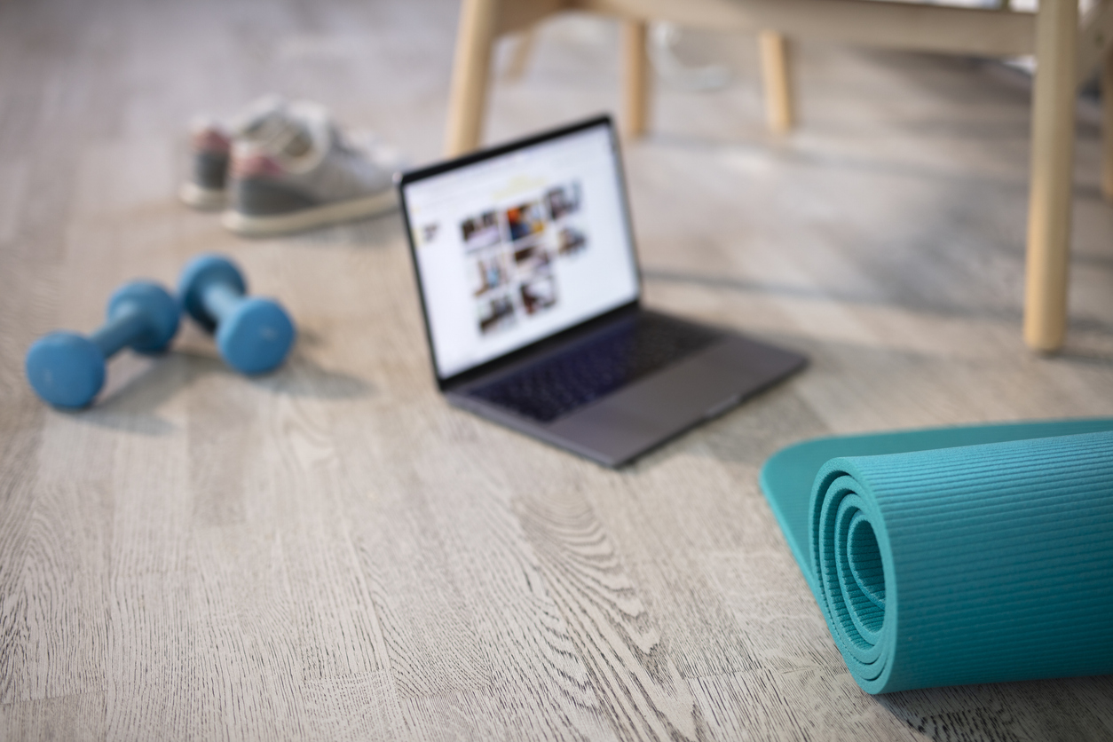 A blue yoga mat, a pair of blue dumbbells, and running shoes rest on a wooden floor near an open laptop displaying various images, suggesting a home workout setup that’s perfect for those looking to save money.