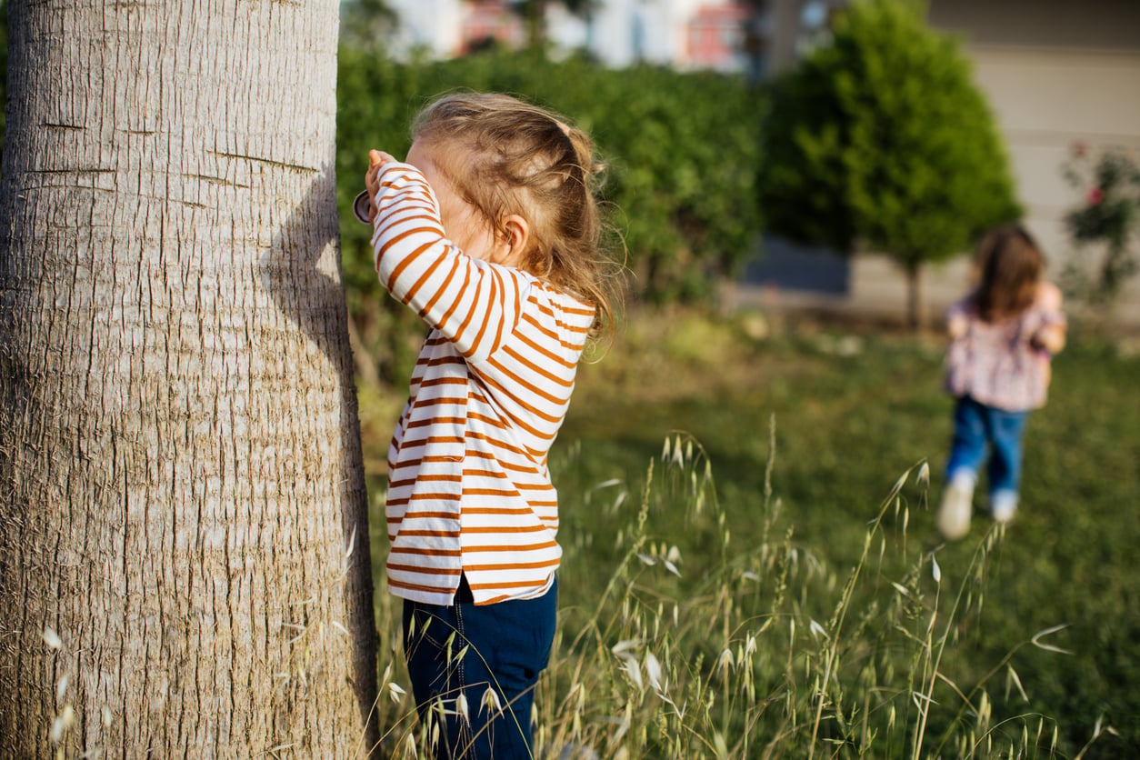 A young child in a striped shirt covers their eyes, standing by a tree, while another child walks away in the grass in the background.