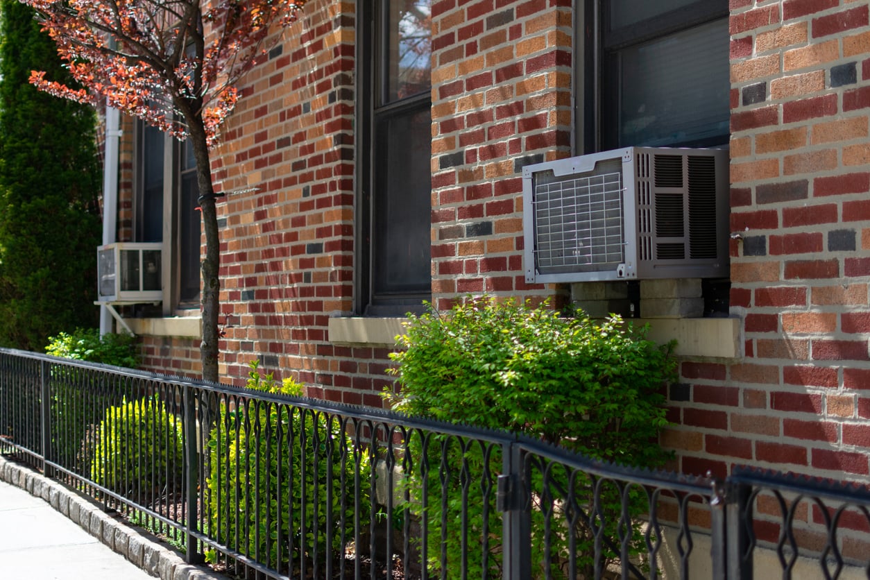 A brick apartment building with black-framed windows, two window air conditioning units, green bushes, a small tree, and a black iron fence along the sidewalk on a sunny day.