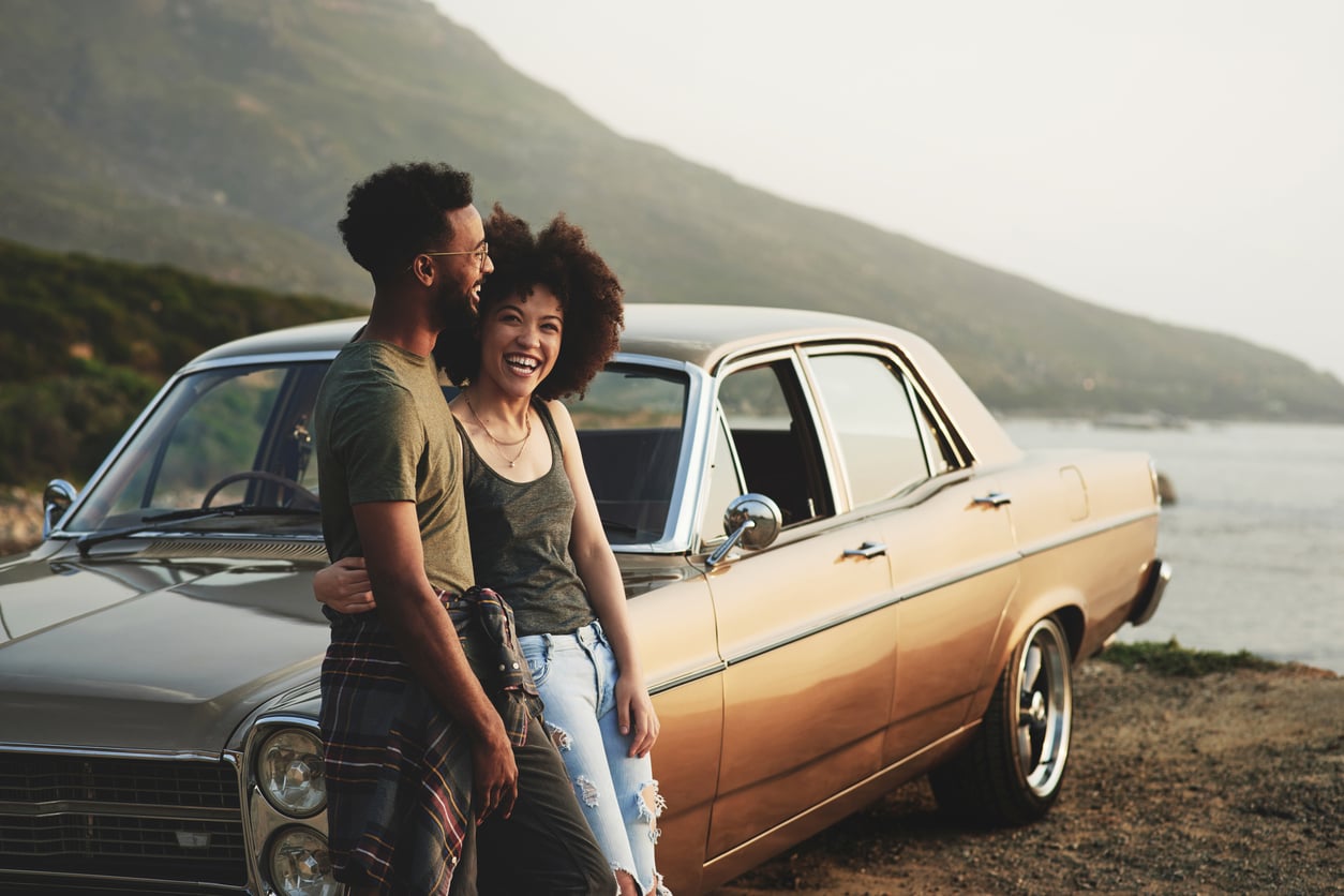 A smiling couple stands by a vintage car parked near a scenic coastal road. The woman sits on the car’s hood while the man stands beside her, perhaps sharing car maintenance hacks as hills and water create a relaxed, joyful atmosphere.