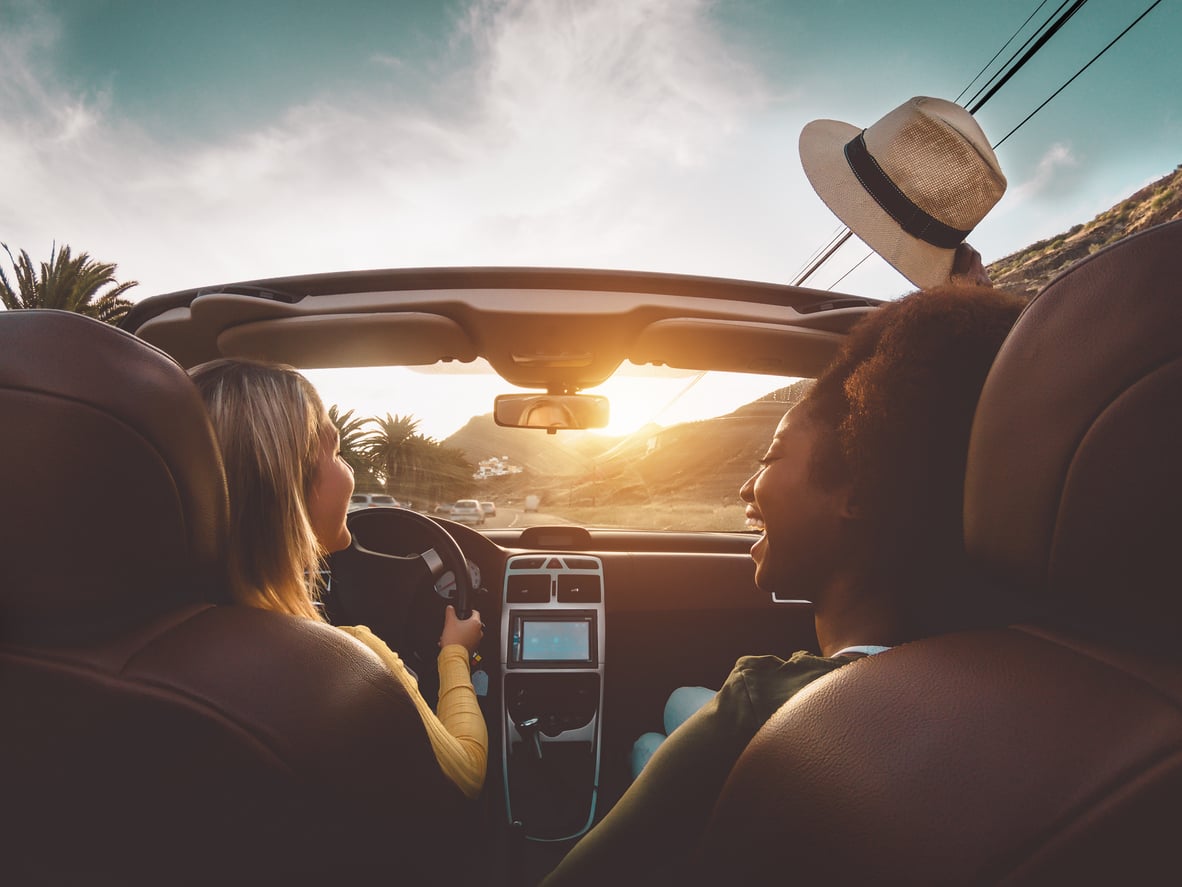 Two people drive a convertible along a scenic road at sunset. One is driving, while the other smiles and wears a hat. Palm trees and hills are visible outside, and the dashboard and car seats are seen from behind.