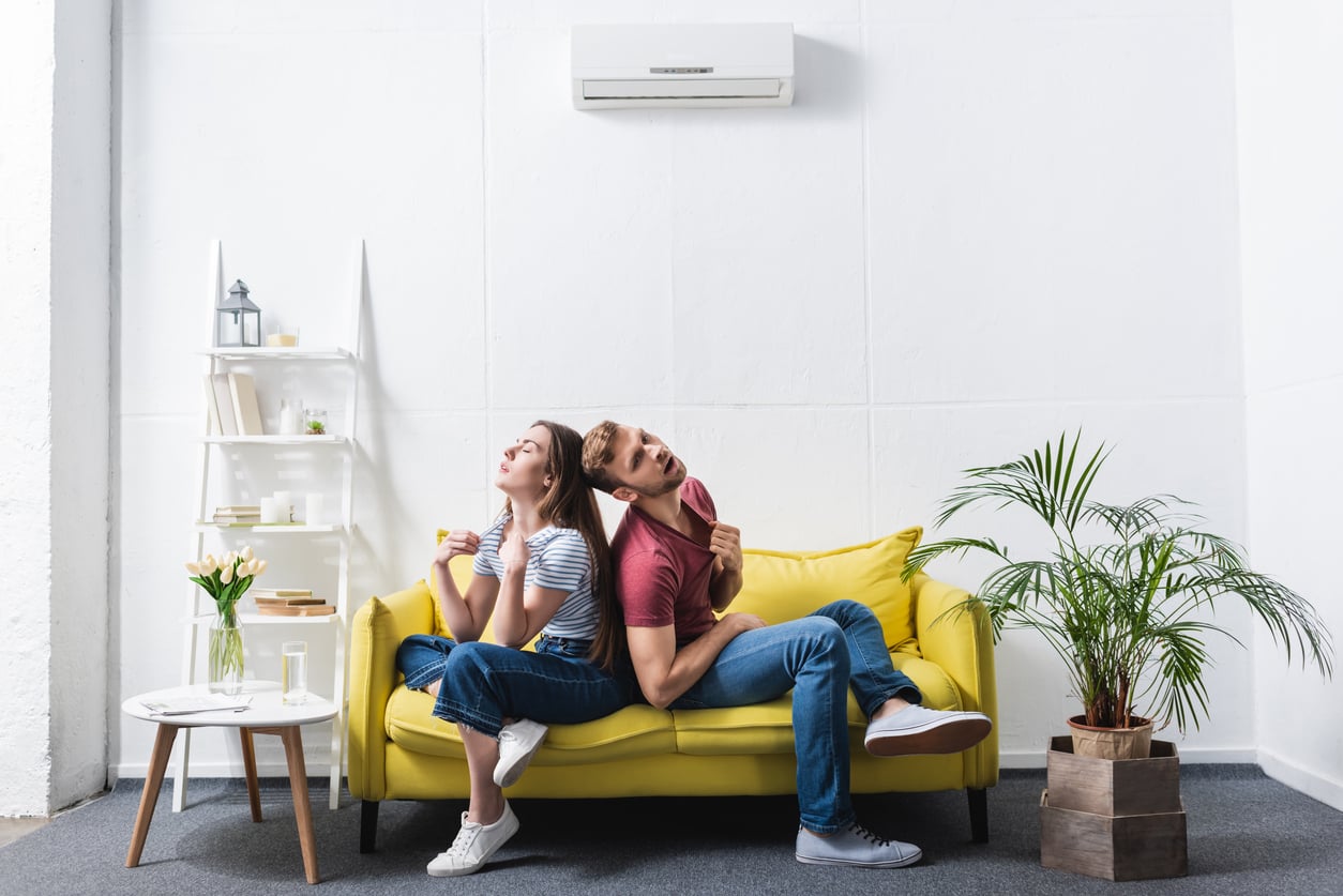 A young couple sits back-to-back on a yellow sofa, both looking away and appearing upset. The room is bright with white walls, a shelf, a round table, and green plants as decor.