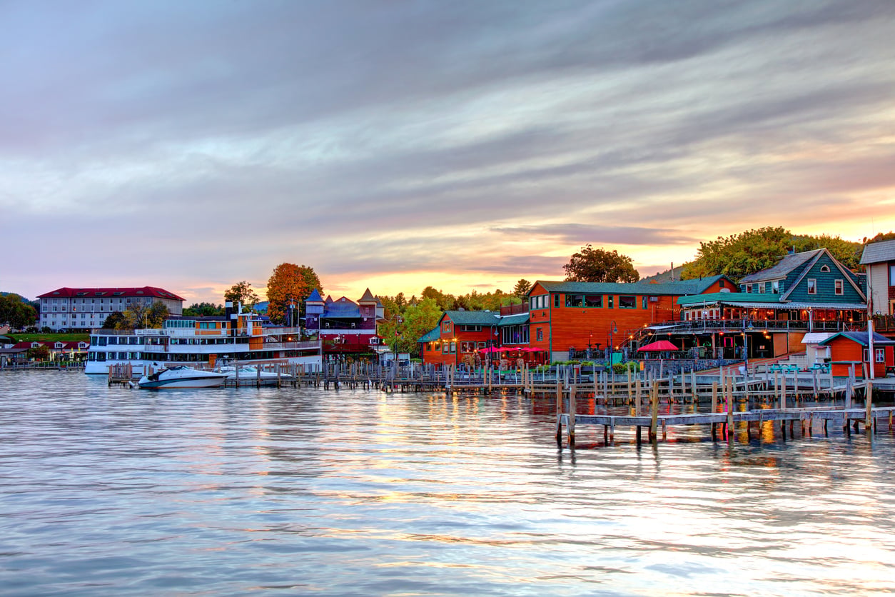 Colorful waterfront buildings and restaurants line a marina at sunset, with boats docked along wooden piers and calm water reflecting the sky and structures.