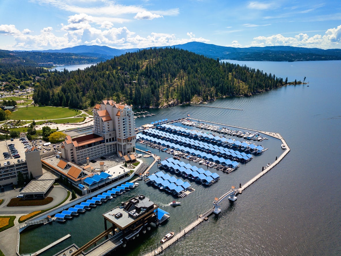 Aerial view of a large marina with covered docks, next to a resort hotel and forested hills by a lake under a partly cloudy sky.