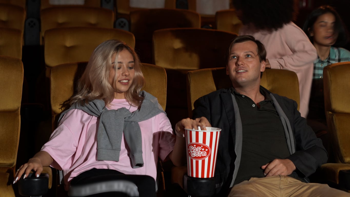 A woman and man sit in a movie theater with brown seats; the man is reaching into a large striped popcorn bucket between them, perhaps trying out some movie theater hacks. Other people are visible in the background.