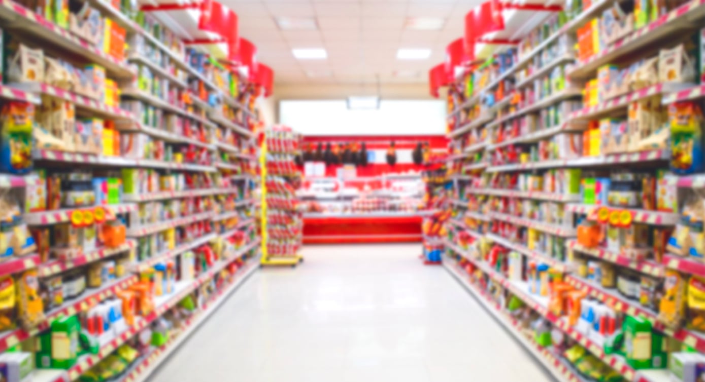 A blurred image of supermarket aisles filled with various colorful products on shelves, leading toward the refrigerated section in the back.