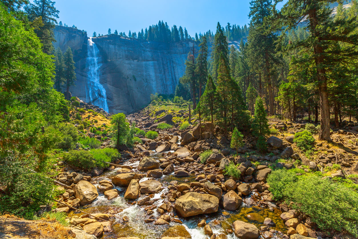 A breathtaking travel destination on the West Coast, this waterfall cascades down a rocky cliff surrounded by tall pine trees, lush greenery, and scattered boulders under a clear blue sky.