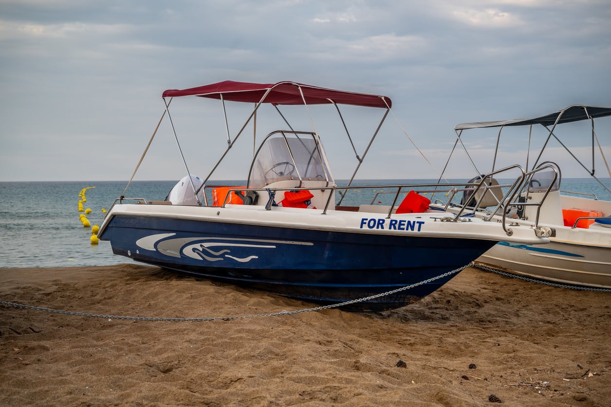 A blue and white motorboat with a red canopy and "FOR RENT" sign is beached on a sandy shore, with safety vests on board—offering an affordable way to enjoy the water without worrying about vacation costs. The sea and a cloudy sky are visible in the background.