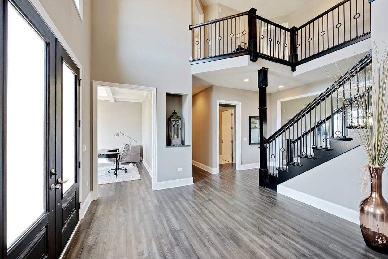 Modern home entryway with dark double doors, gray wood flooring, open staircase with black railing, and a hallway leading to an office with a desk and chair. Large vase with tall branches is placed near the stairs.