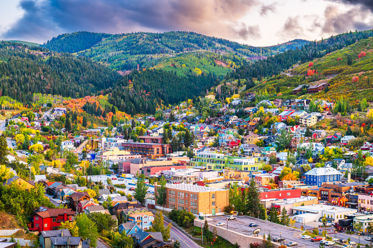 Colorful buildings and houses sit nestled in a green, hilly landscape under a partly cloudy sky, with forests and mountains in the background—making this picturesque scene one of the most charming travel destinations.