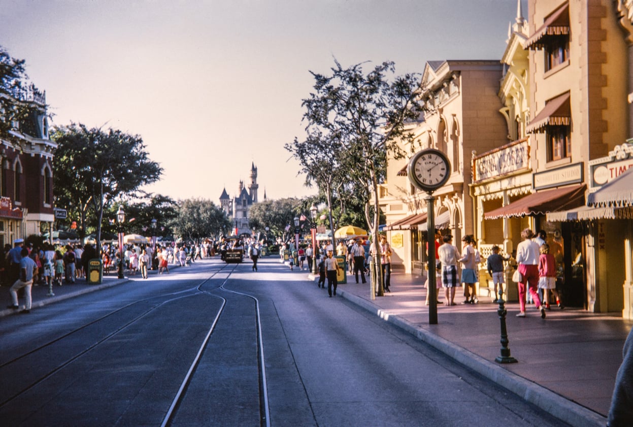 A vintage photo of Disneyland’s Main Street, USA, with people walking, shops on either side, a street clock, and Sleeping Beauty Castle visible in the distance under a clear sky—a nostalgic glimpse into one of the world’s most beloved and surprisingly affordable travel destinations.