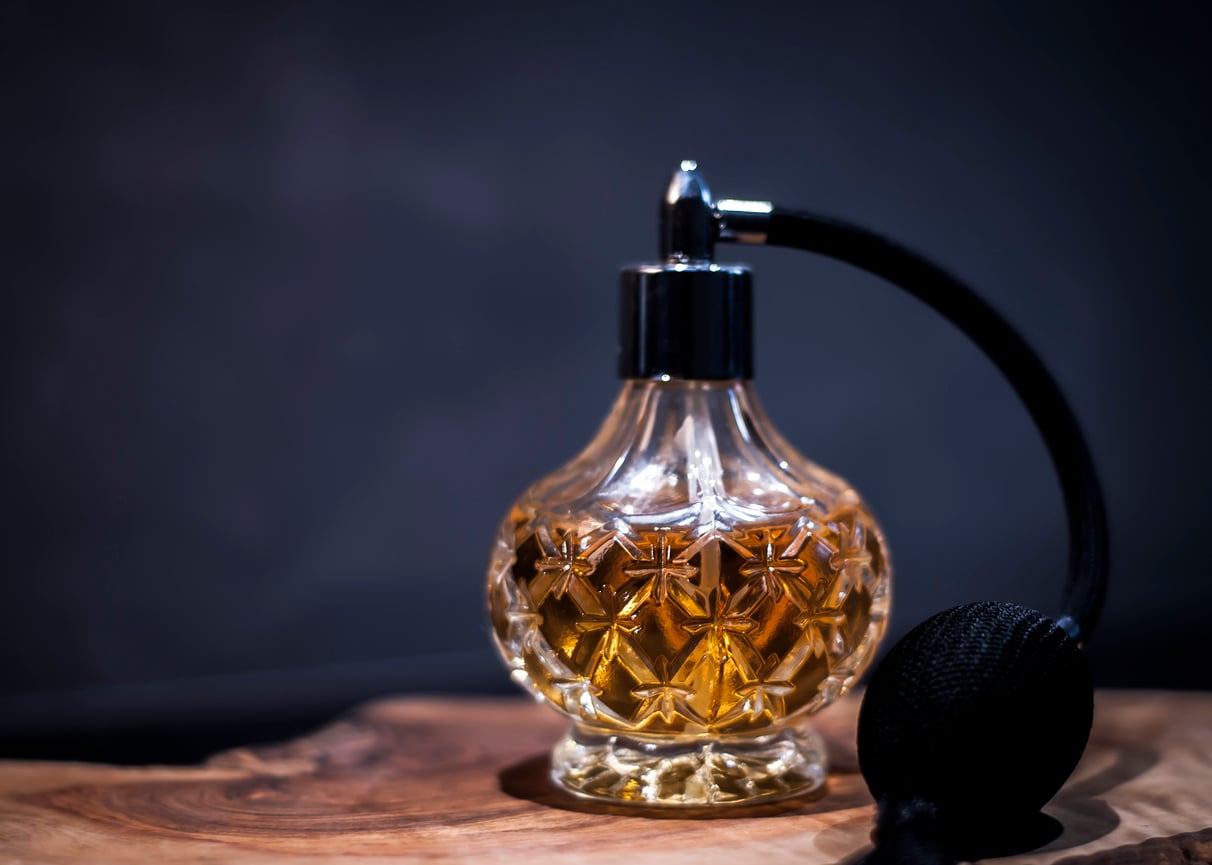 A vintage glass perfume bottle with an ornate pattern holds golden liquid, evoking memories of the signature scents favored by middle-class moms in the 80s and 90s. It has a black atomizer bulb and sits on a wooden surface against a dark, blurred background.
