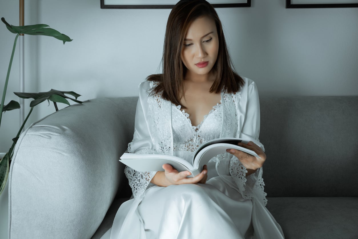 A woman in a white lace robe sits on a gray sofa reading a book, with a plant and framed pictures in the background.
