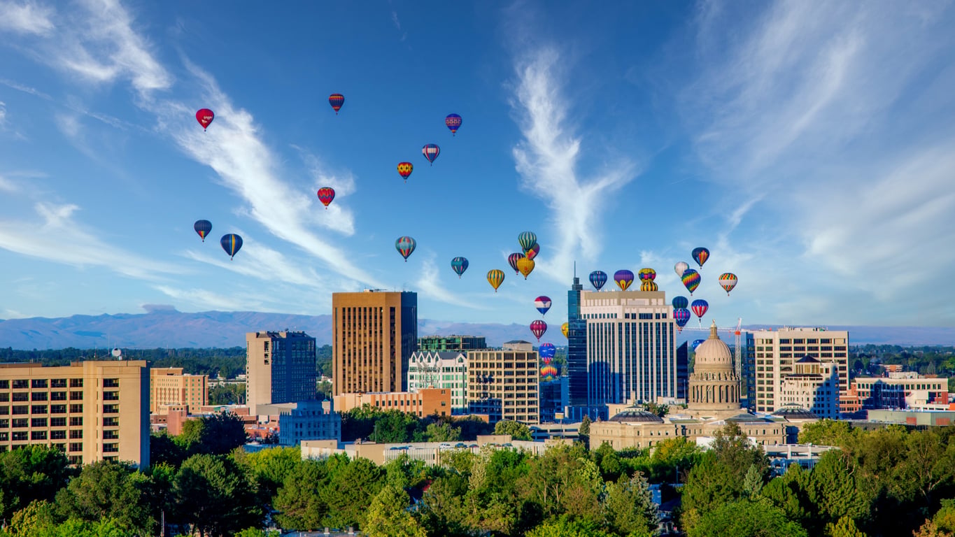 Hot air balloons float above a city with tall buildings and a domed capitol, set against a blue sky with wispy clouds and distant mountains, making it one of the most enchanting travel destinations for adventure seekers and dreamers alike.