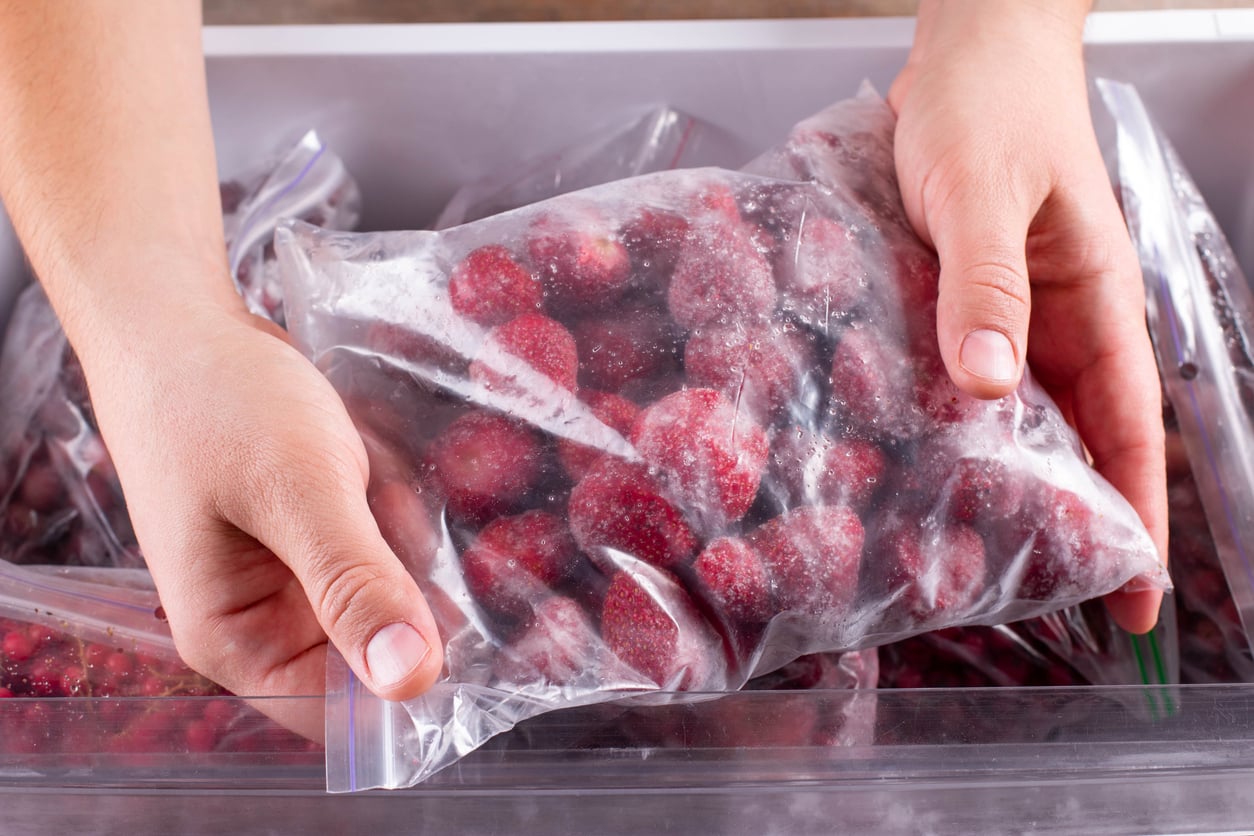 Hands holding a clear plastic bag filled with frozen strawberries, with more bags of frozen fruit visible in a freezer drawer below.