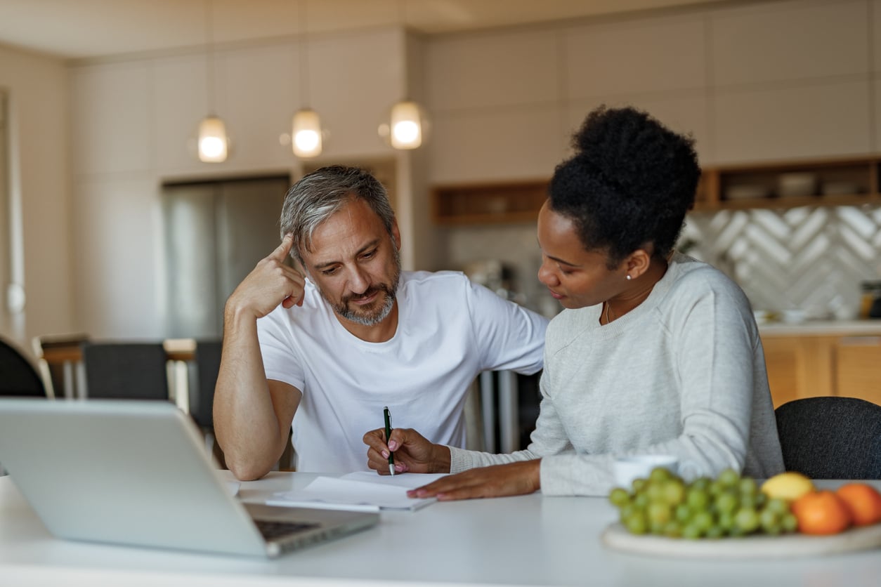A man and woman sit at a kitchen table, looking at paperwork together—perhaps reviewing hotel booking tips for an upcoming trip. The man is holding a pen, while the woman points at the paper, offering her thoughts. A laptop and fruit are on the table.