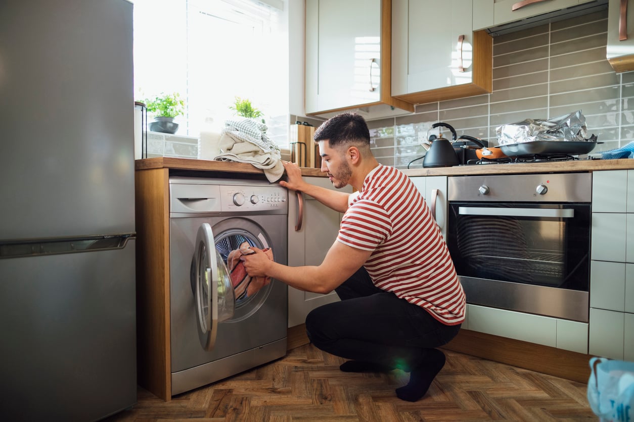 A man in a red and white striped shirt crouches in a modern kitchen, loading clothes into a front-loading washing machine next to an oven and a sink, efficiently managing household expenses.