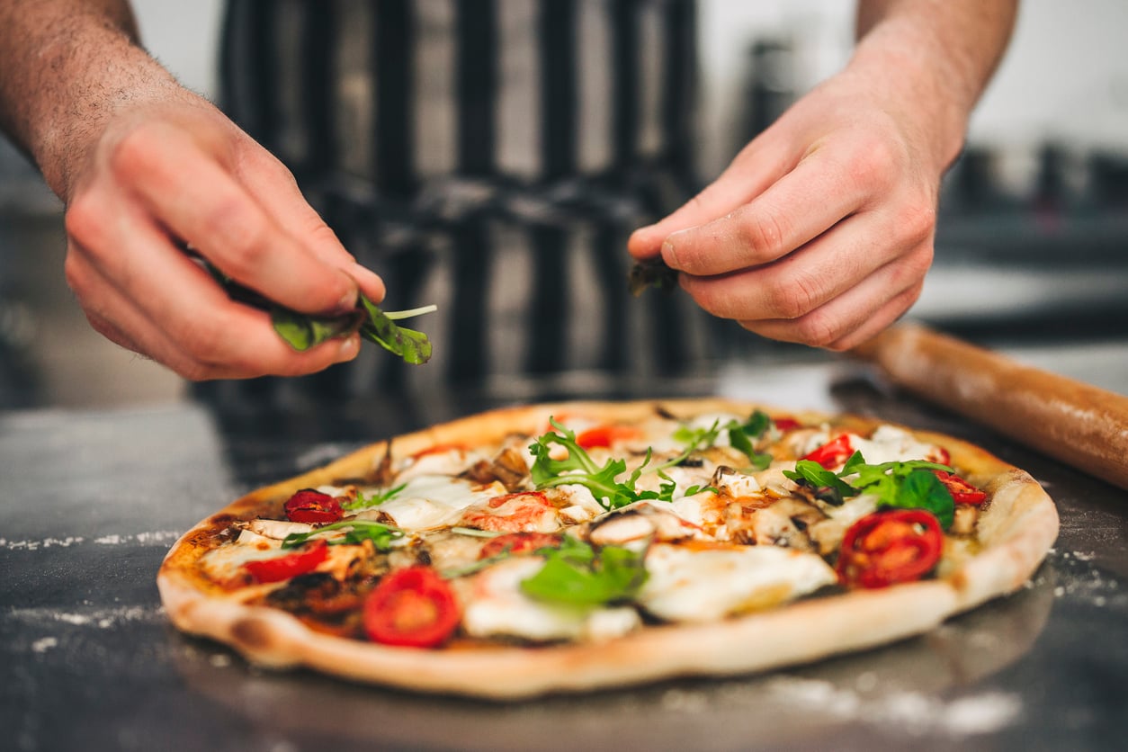 A person adds fresh arugula to a homemade pizza topped with tomatoes, cheese, and vegetables on a kitchen counter.
