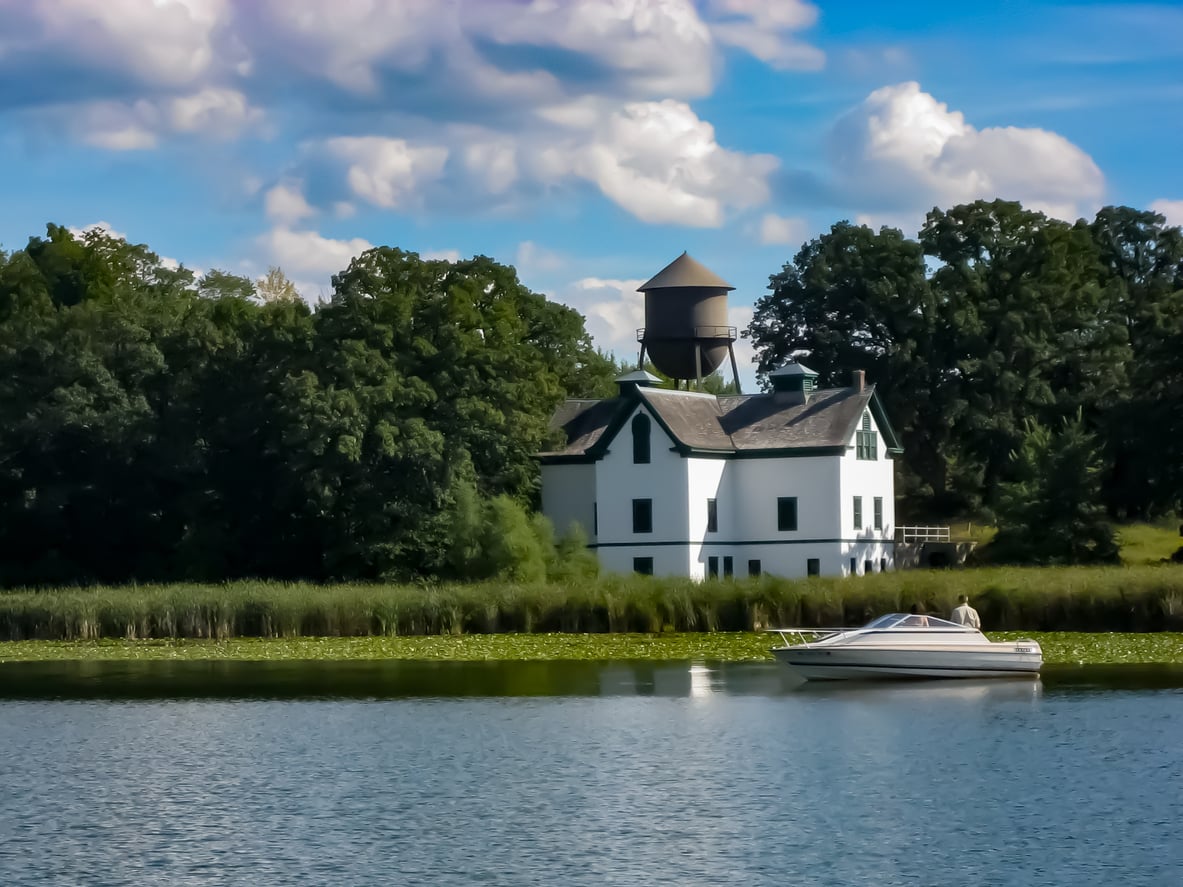 A white house with a water tower stands by a wooded shoreline. A small motorboat floats on the blue lake in the foreground under a partly cloudy sky.