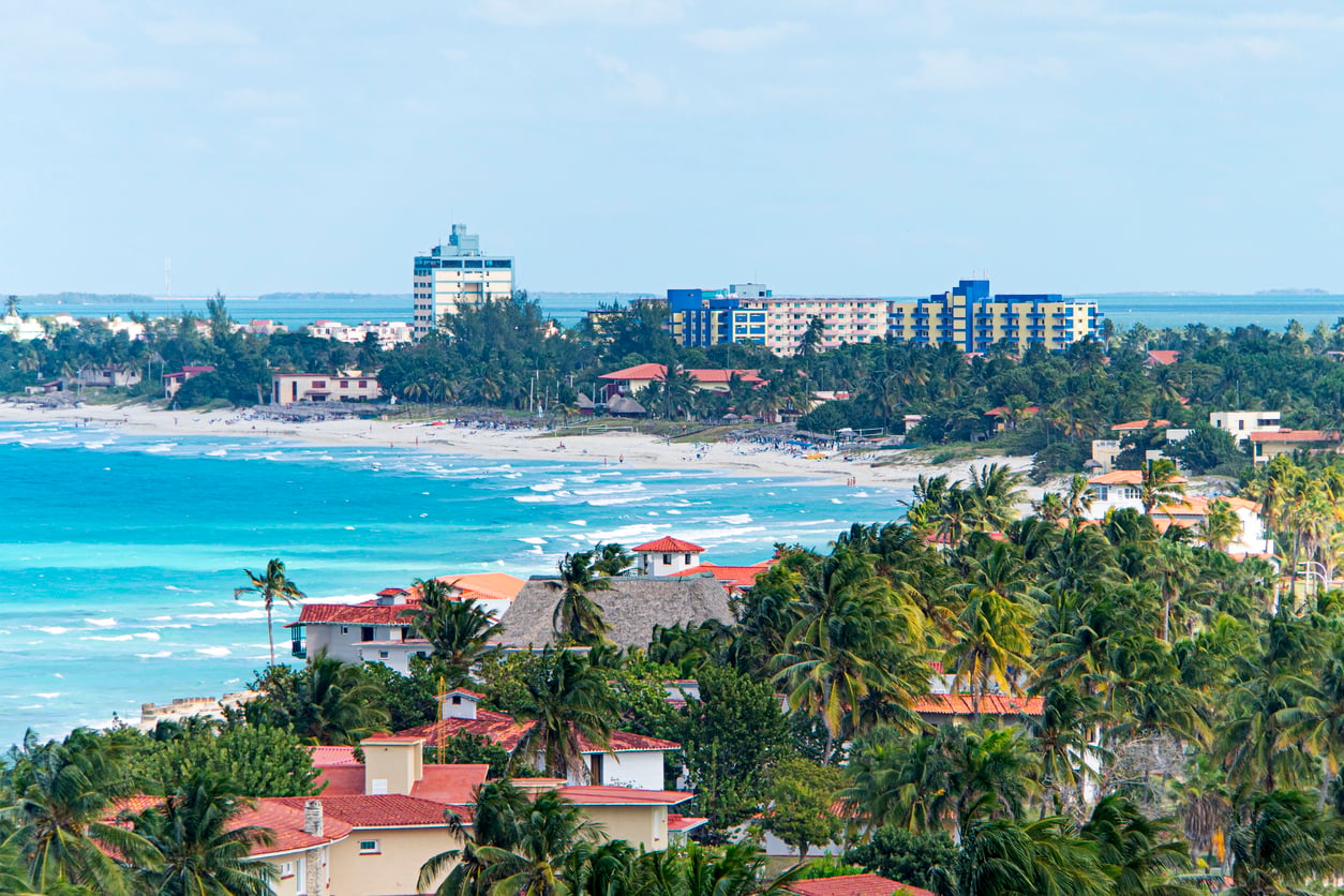 A coastal city with palm trees, colorful buildings, and sandy beaches along turquoise waves—one of the most inviting travel destinations. People are visible on the beach, while mid-rise hotels and apartments line the shoreline under a partly cloudy sky.