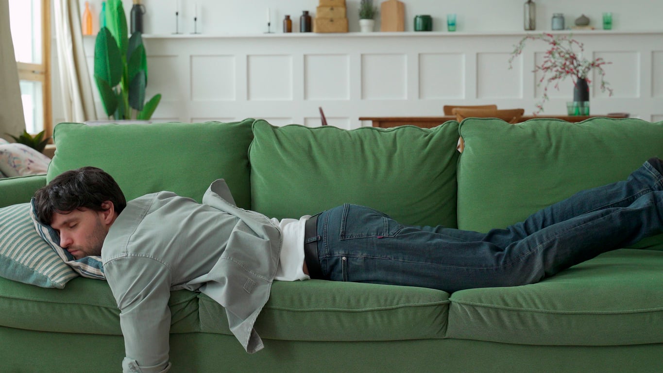 A man, perhaps embodying traditional Baby Boomer beliefs about relaxation and leisure, is lying face down on a green couch, appearing to be asleep. He is wearing jeans and a gray shirt. The background shows a table, chairs, plants, and decorative items on a shelf.
