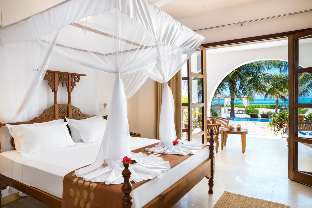 A bright bedroom with a canopy bed, white linens, and towels shaped like swans. Glass doors open to a patio with palm trees, a pool, and a view of the ocean.