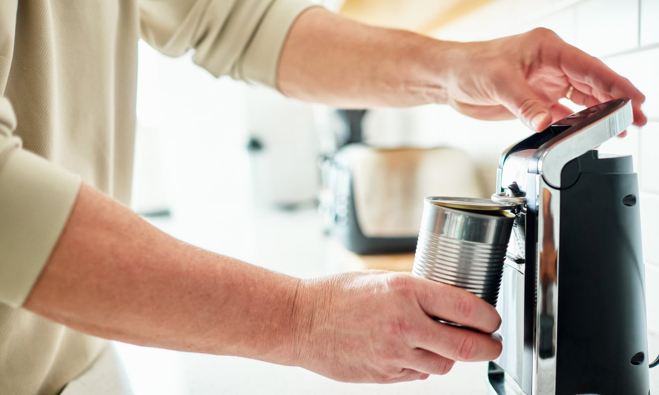 A person using an electric can opener—one of the essential kitchen gadgets—to open a metal can in a bright kitchen, with a toaster visible in the background.
